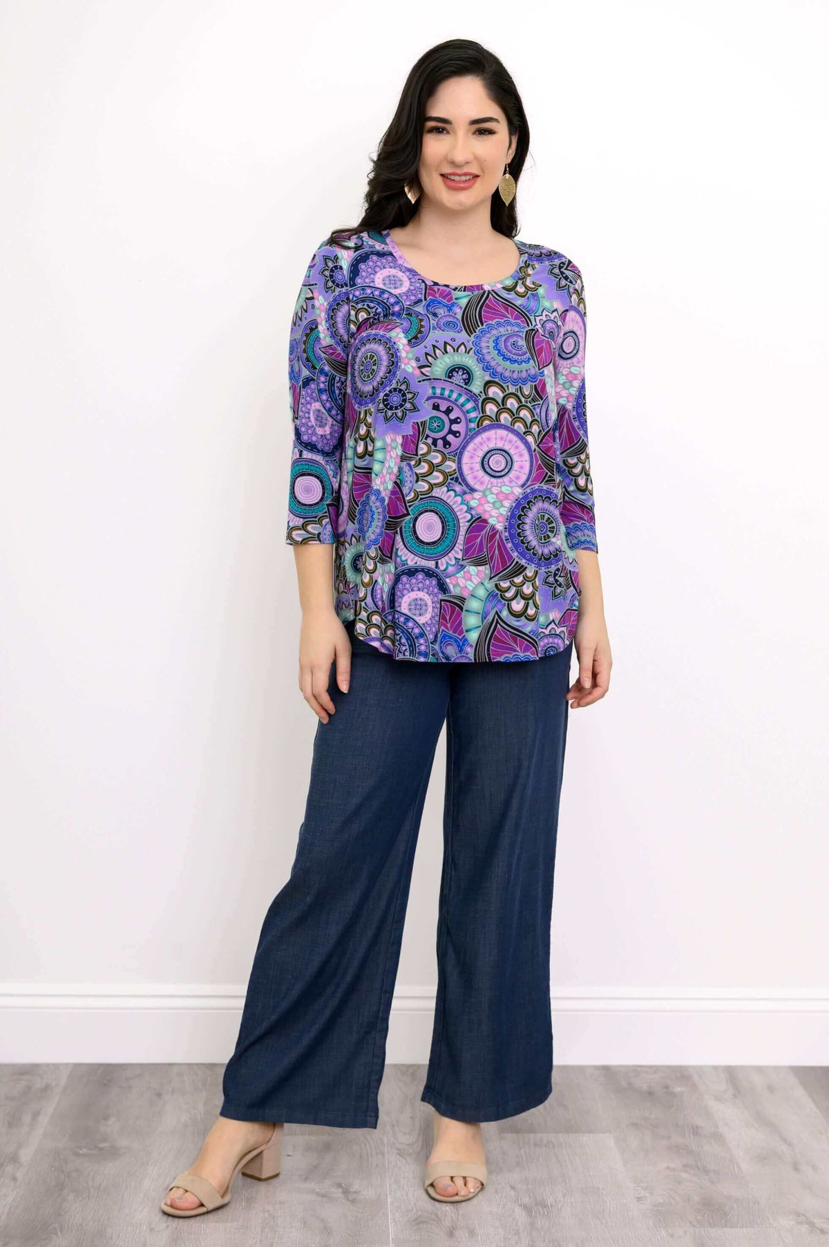 Full body front view of woman  smiling, wearing  scoop neck purple patterned top and denim pants