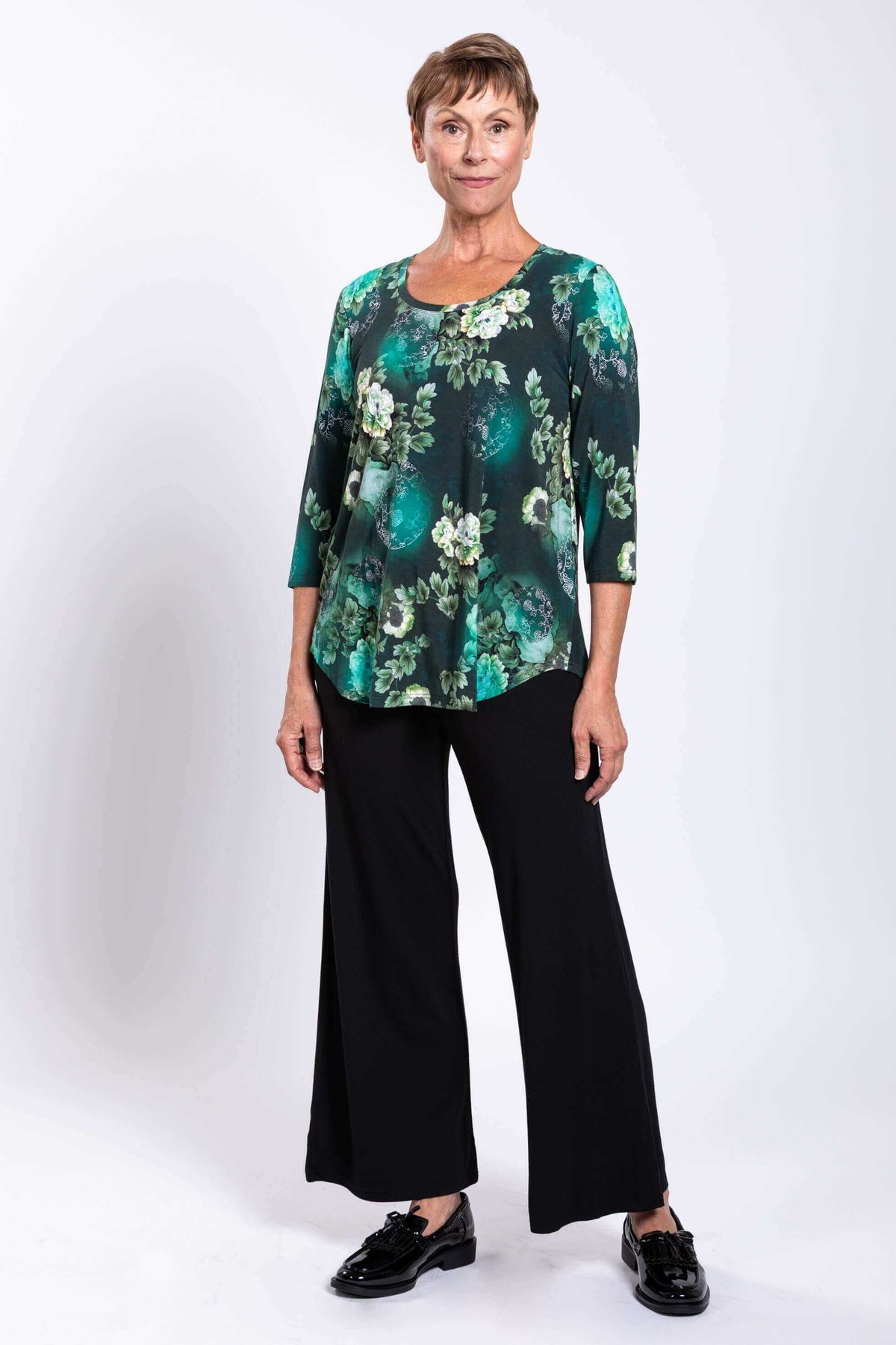 Three-quarter front view of woman in green floral 3/4 sleeve bamboo top and black pants