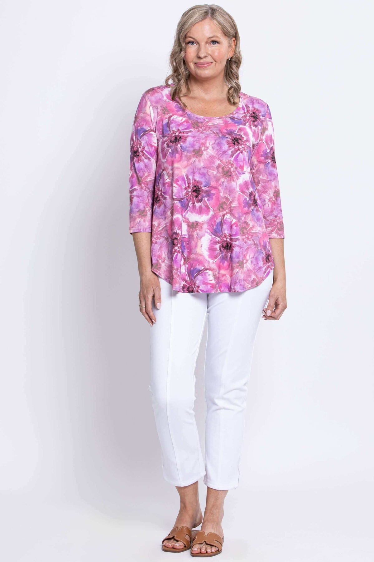 Woman modeling with her arms relaxed in a pink floral top, white pants, and tan colored sandals