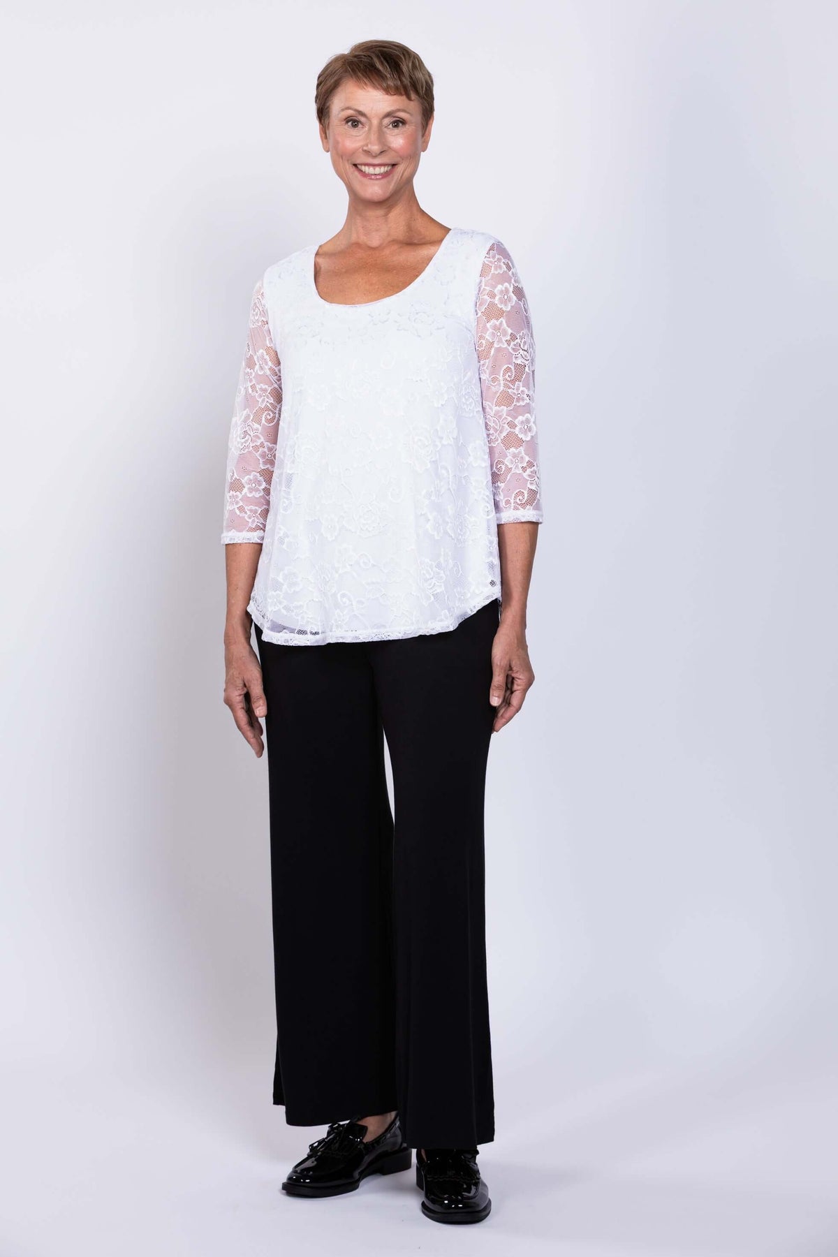 Angled front view of a woman in a white lace overlay top with 3/4 sleeves and black pants