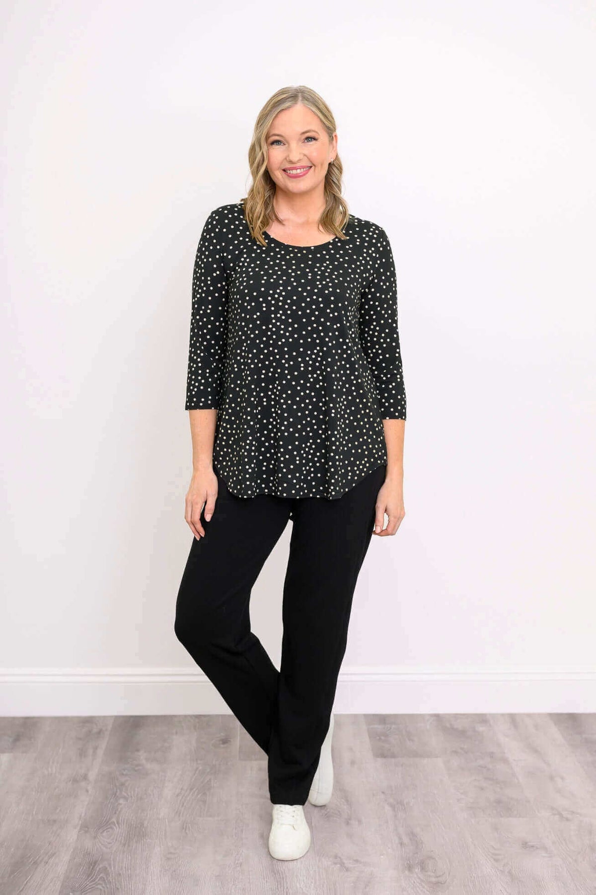 Smiling woman in black polka dot 3/4 sleeve top and black pants with white sneakers