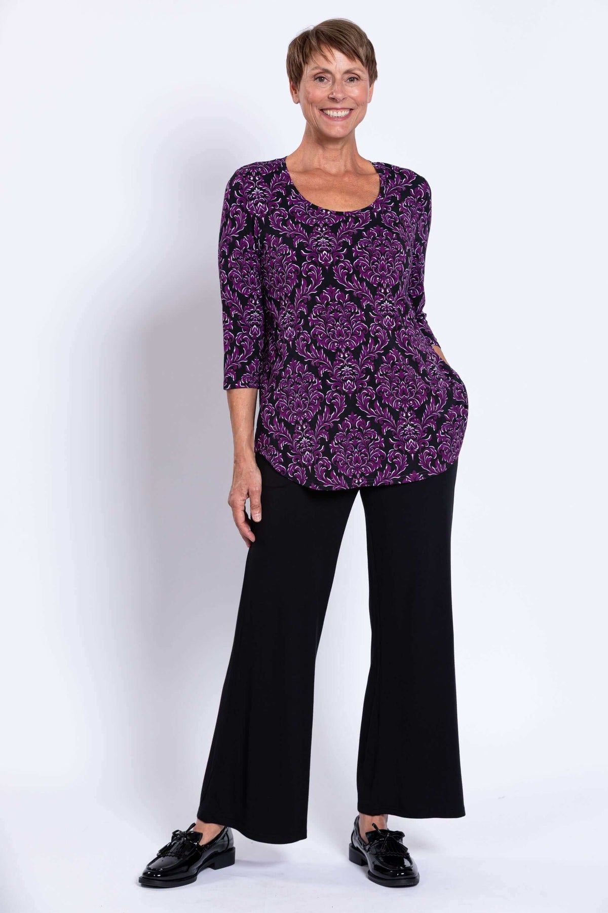 Woman modeling a purple damask Jazz 3/4 sleeve top with black pants, hand in pocket, front view