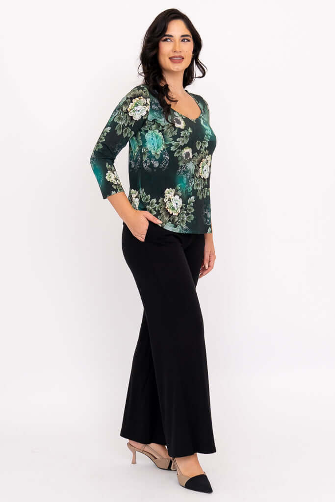 Woman in green floral sweetheart top and black pants, angled side view with hand in pocket