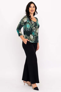 Woman in green floral sweetheart top and black pants, angled side view with hand in pocket