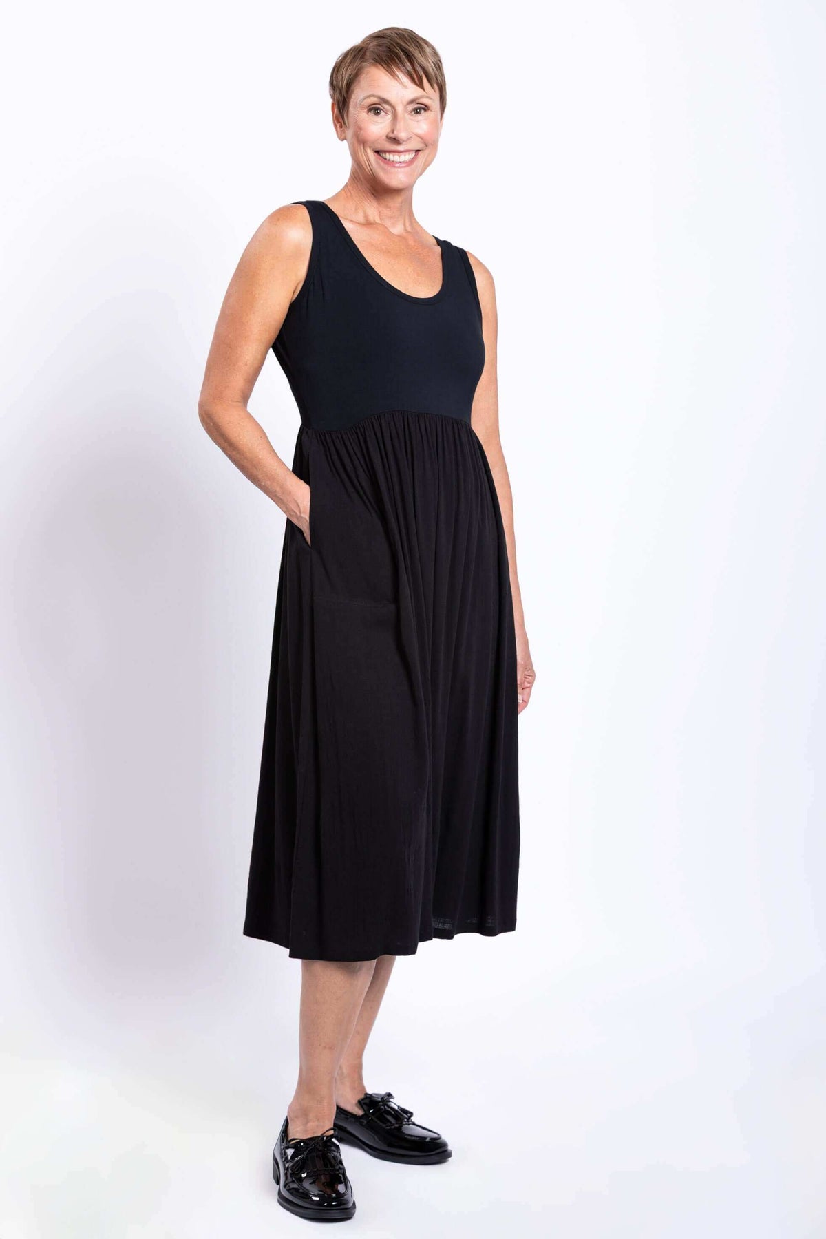 Angled side view of a woman in a black sleeveless empire waist midi dress, hand in pocket
