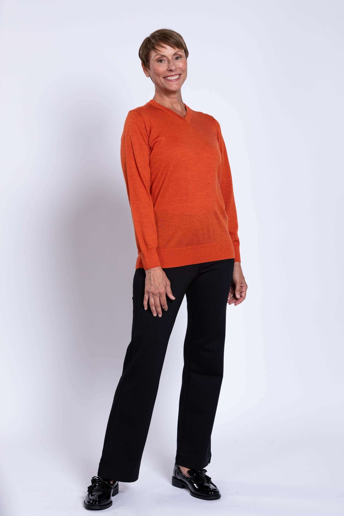 Woman smiling, wearing a tangerine V-neck merino wool sweater and black pants, angled pose.