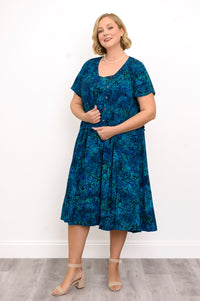 Woman wearing a blue patterned dress and blue patterned blouse standing against a white wall, front view