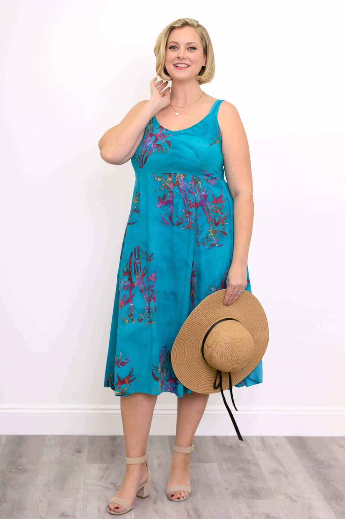 Woman in an aqua blue floral midi dress holding a straw sunhat and smiling