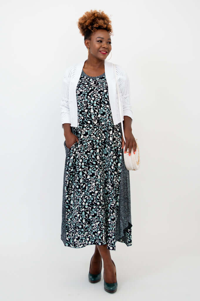 Model with white cardigan over black and blue patterned tank dress, holding a clutch