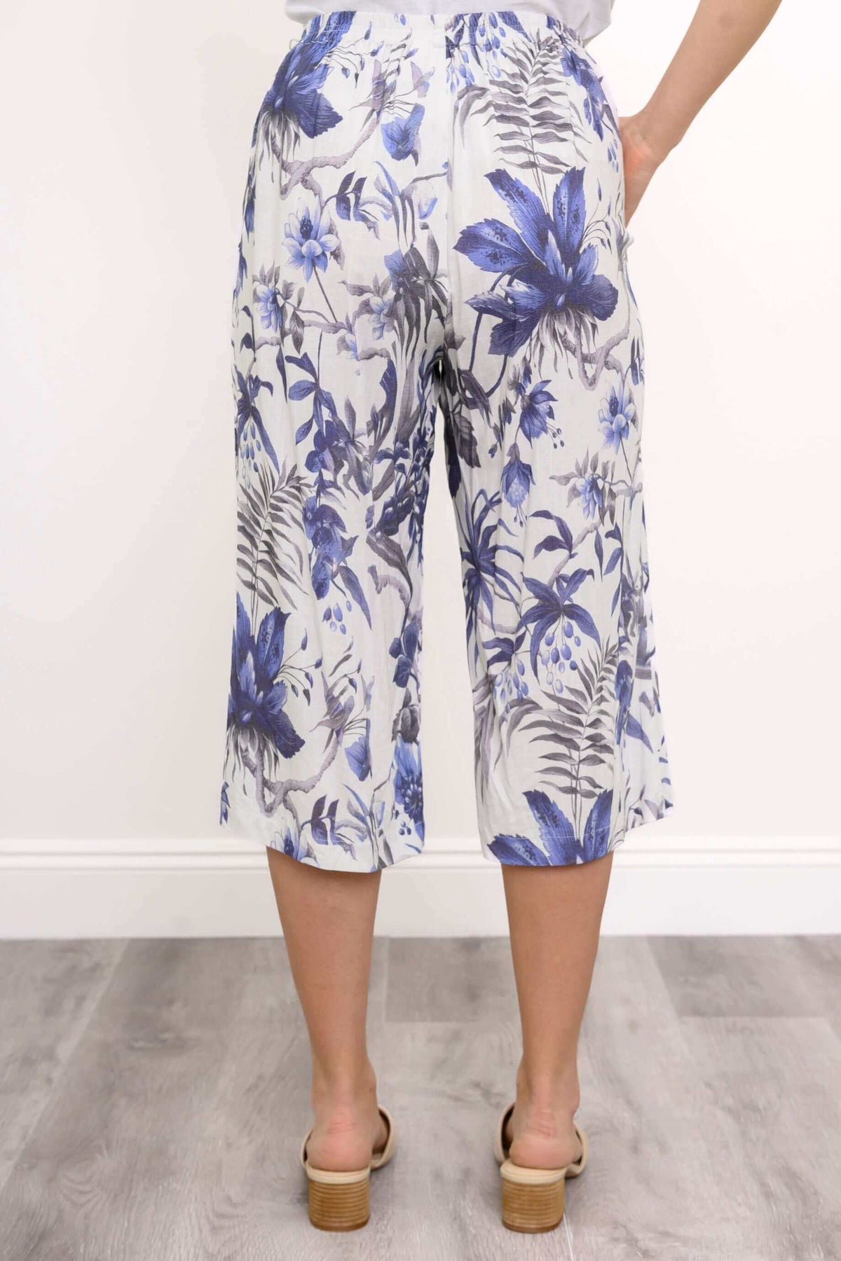 Back close-up of woman with her hand in her pocket wearing a paid of linen white capri pants with blue florals