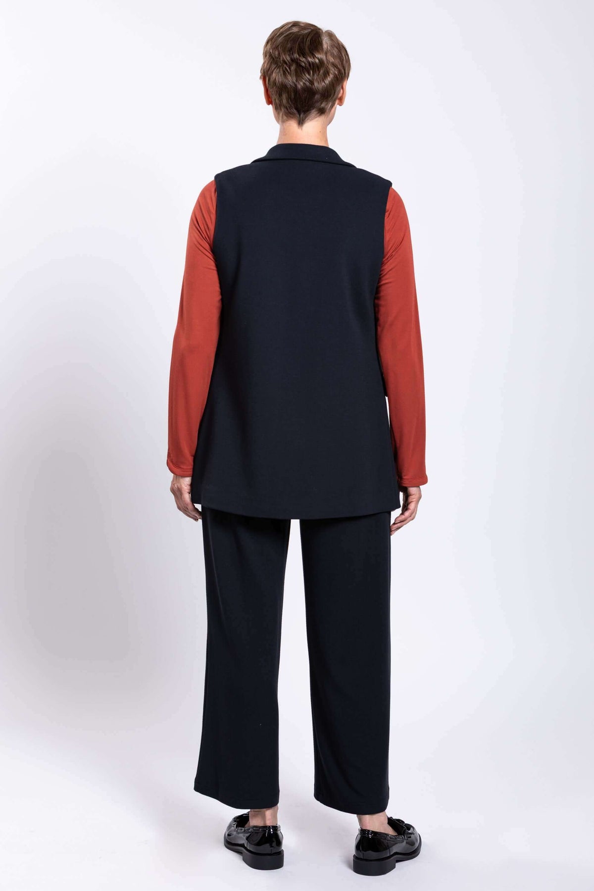 Back view of model in black open vest, matching pants, and rust colored long sleeve shirt