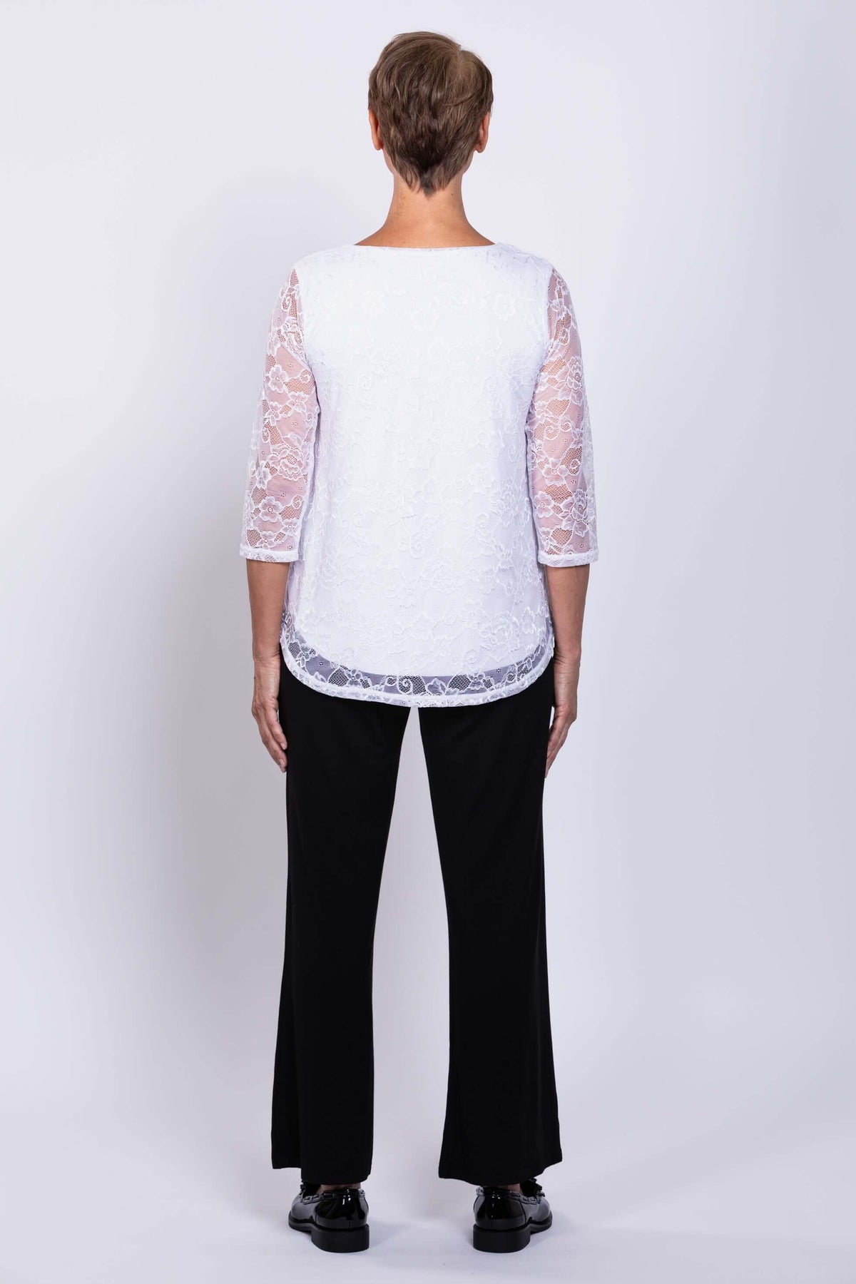Back view of a woman wearing a white lace overlay top with 3/4 sleeves and black pants