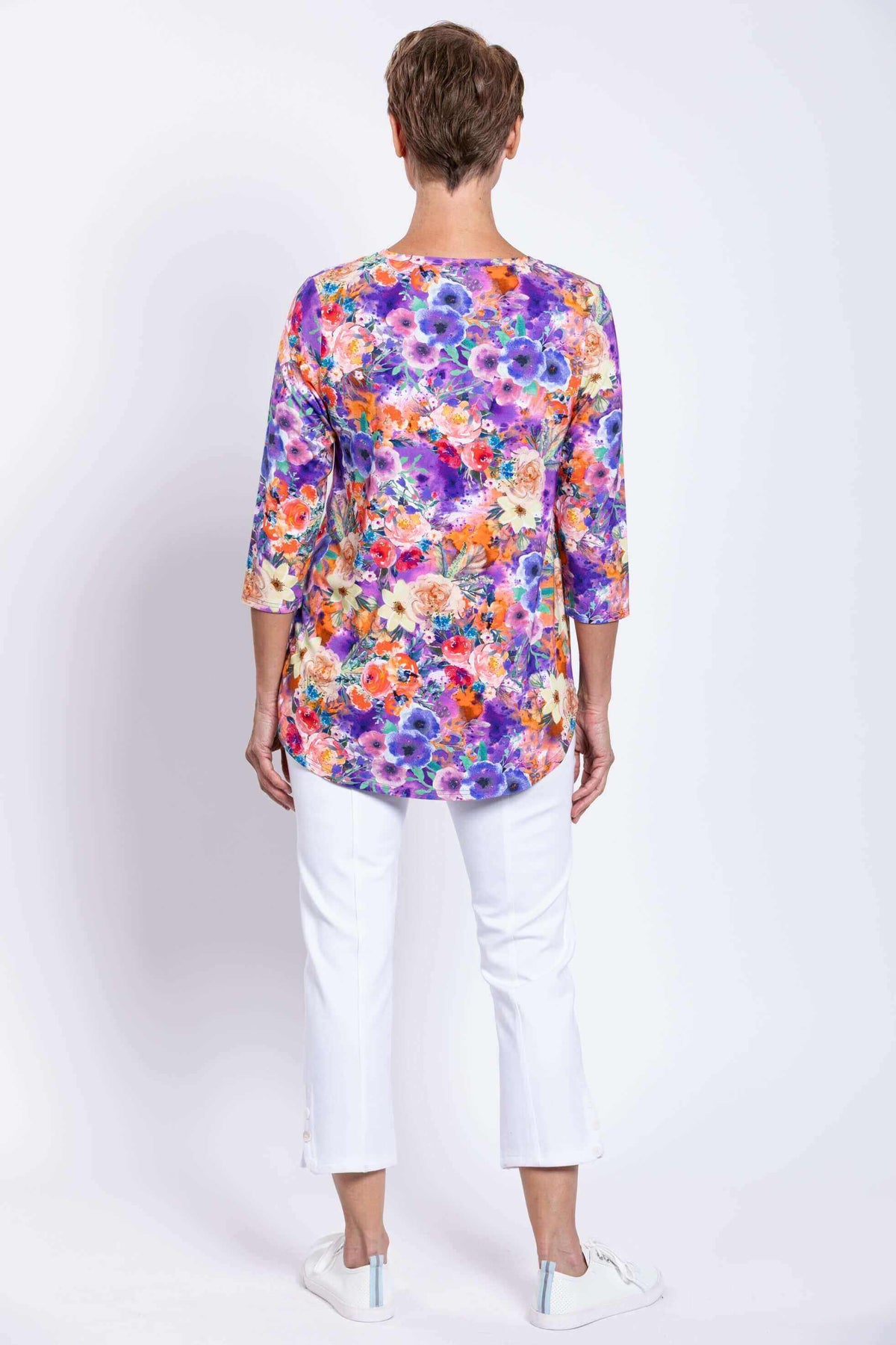 Back view of woman wearing bright floral top and white pants against a white background