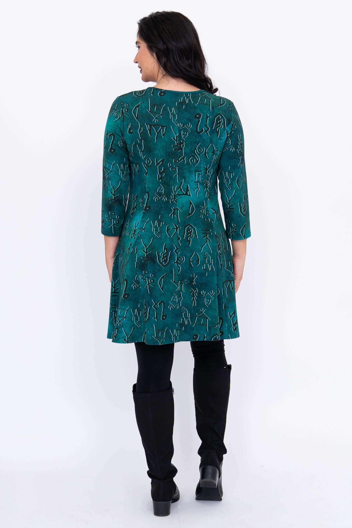Back view of a woman in a teal tunic with petroglyph pattern and long black boots