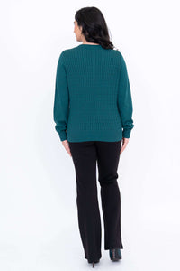 Back view of a woman in a teal cable knit sweater with ribbed hem, wearing black pants