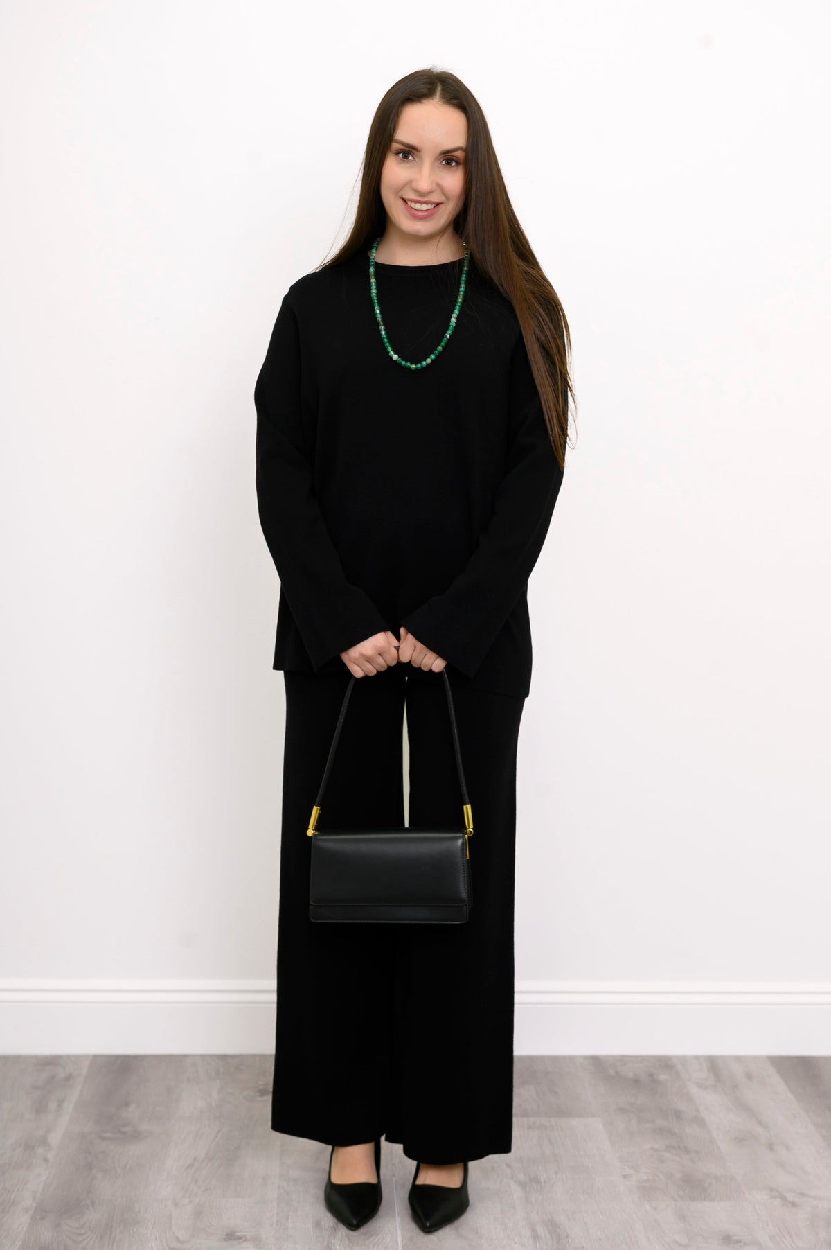 Smiling woman styling black straight-leg trousers with a green statement necklace and a matching black purse.