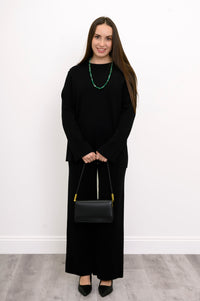 Smiling woman styling black straight-leg trousers with a green statement necklace and a matching black purse.