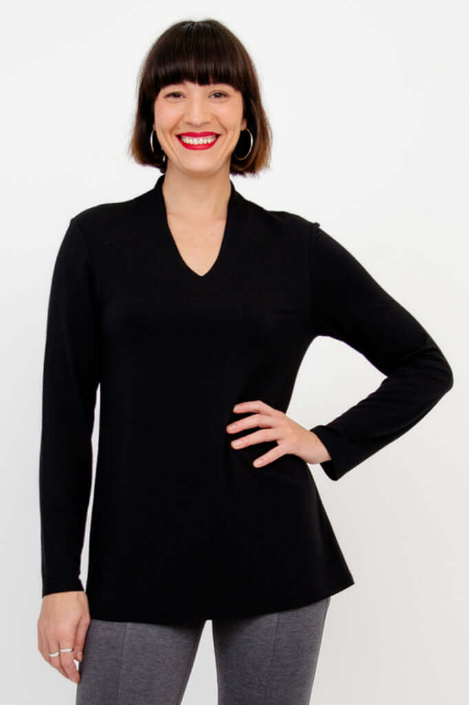 Woman wearing a black long sleeve v-neck top and grey pants, smiling with hand on hip