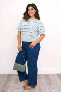 Woman in light blue eyelet stripe sweater and jeans holding a gray purse, angled side view
