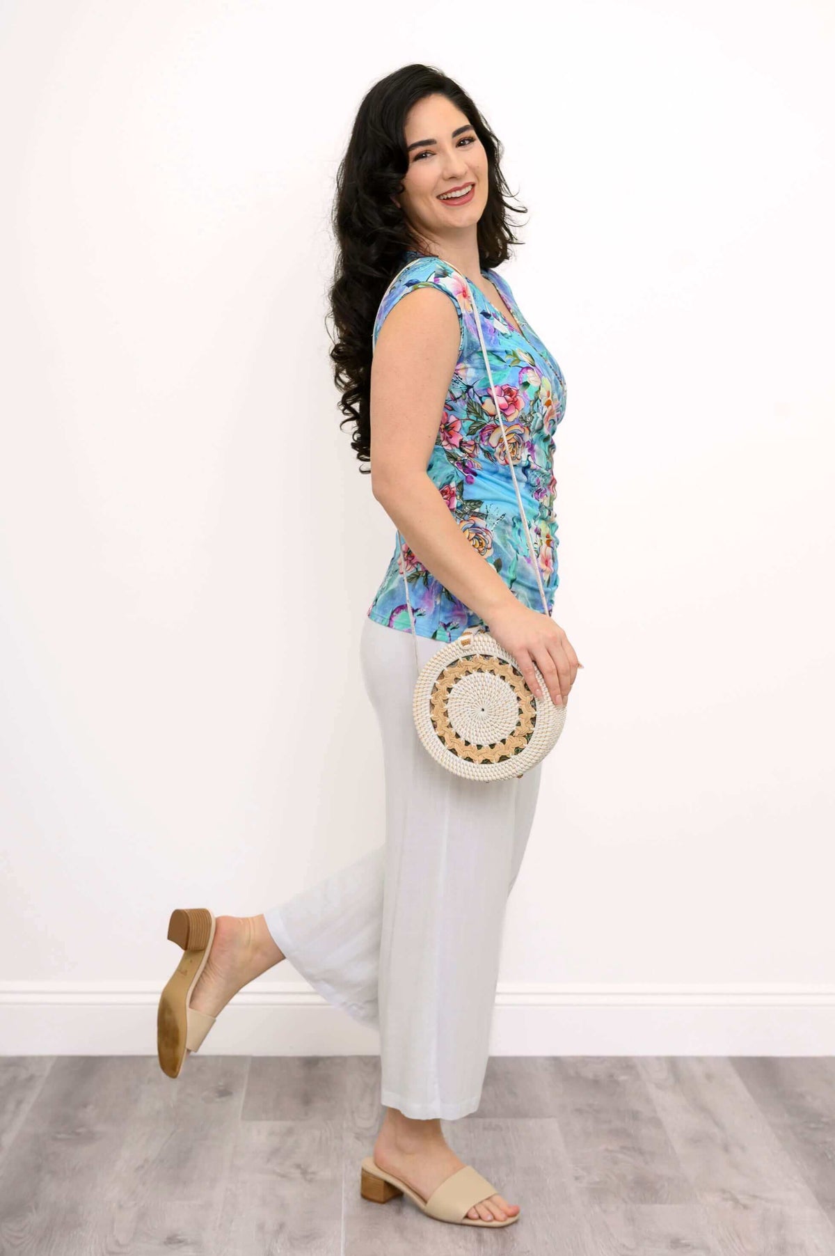 Woman in blue floral Nova Tank, cap sleeves, with white pants and a round woven bag, side view