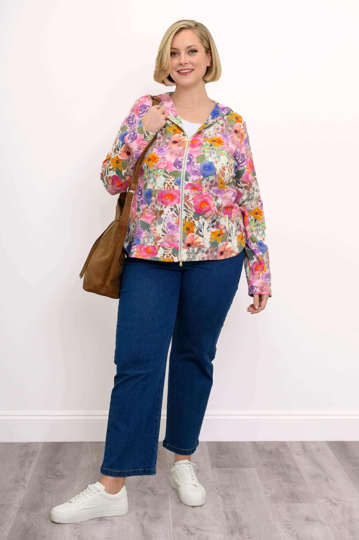 Woman holding a brown bag, dressed in a pink floral hoodie jacket with jeans and white sneakers
