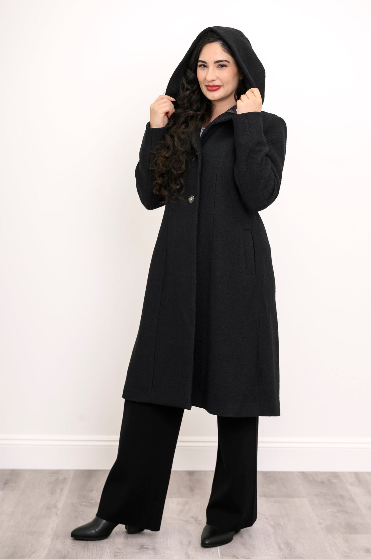 Woman posing with her coat hood on in a black loose-fitting coat and black pants
