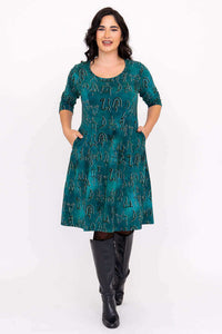 Woman wearing teal petroglyph dress with hands in side pockets, facing forward and smiling