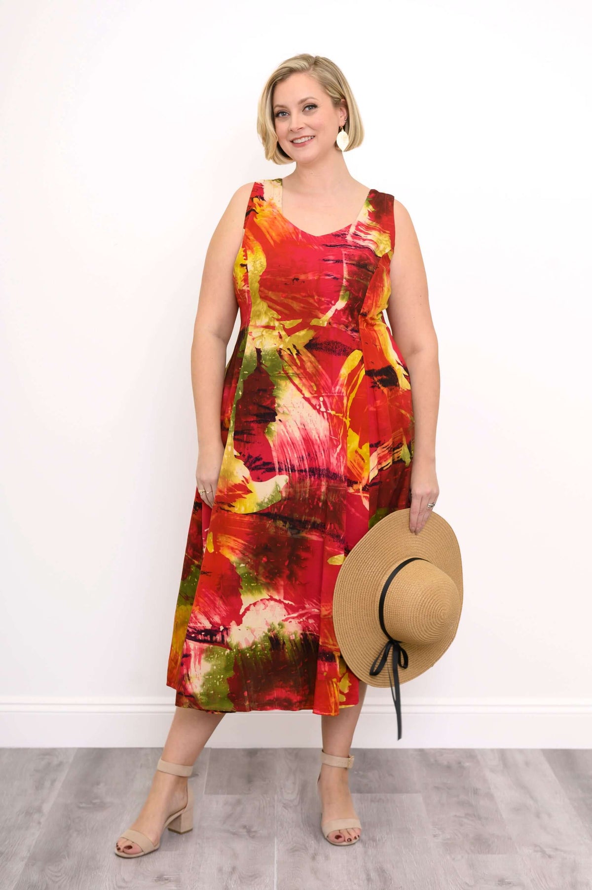 Woman in a vibrant abstract print sleeveless midi dress, smiling and holding a straw hat by her side