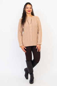 Woman stands with relaxed pose in beige cable knit sweater and black boots