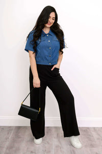 Woman posing with a black handbag in a blue cropped denim jacket and black pants