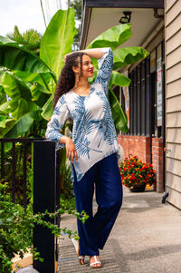 Woman outdoors in a white top with blue palm print and navy pants, standing by plants