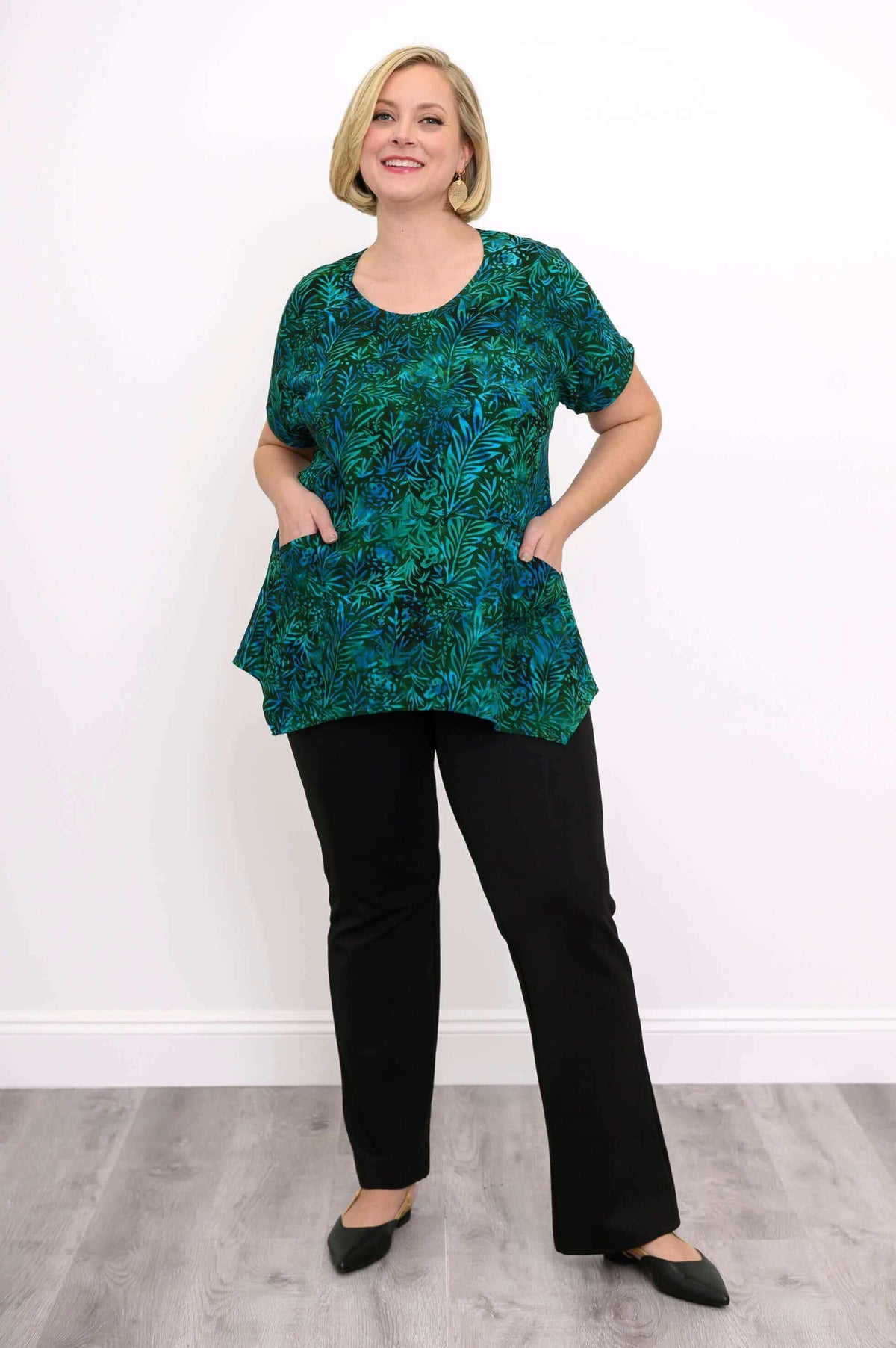 Woman posing with hands in pockets of green and blue leaf print tunic top, wearing black pants