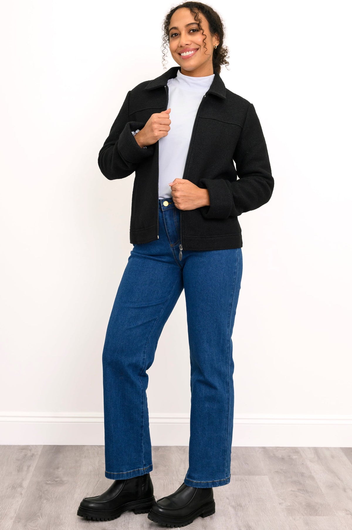 Woman wearing a black jacket, white shirt, blue jeans, and black shoes on a white background, front view
