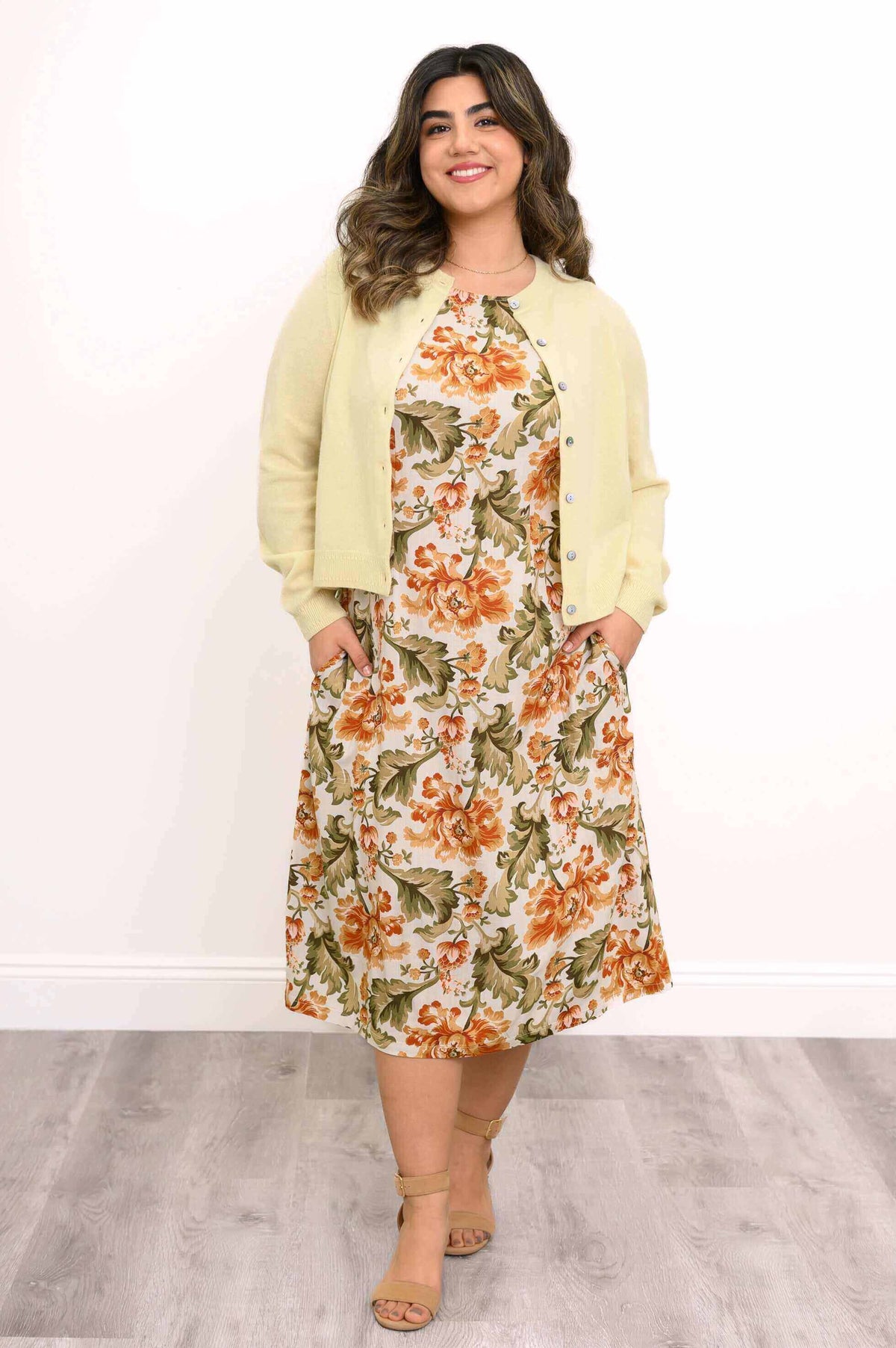 Woman wearing floral dress with yellow cardigan and beige heels, hands in pockets, smiling
