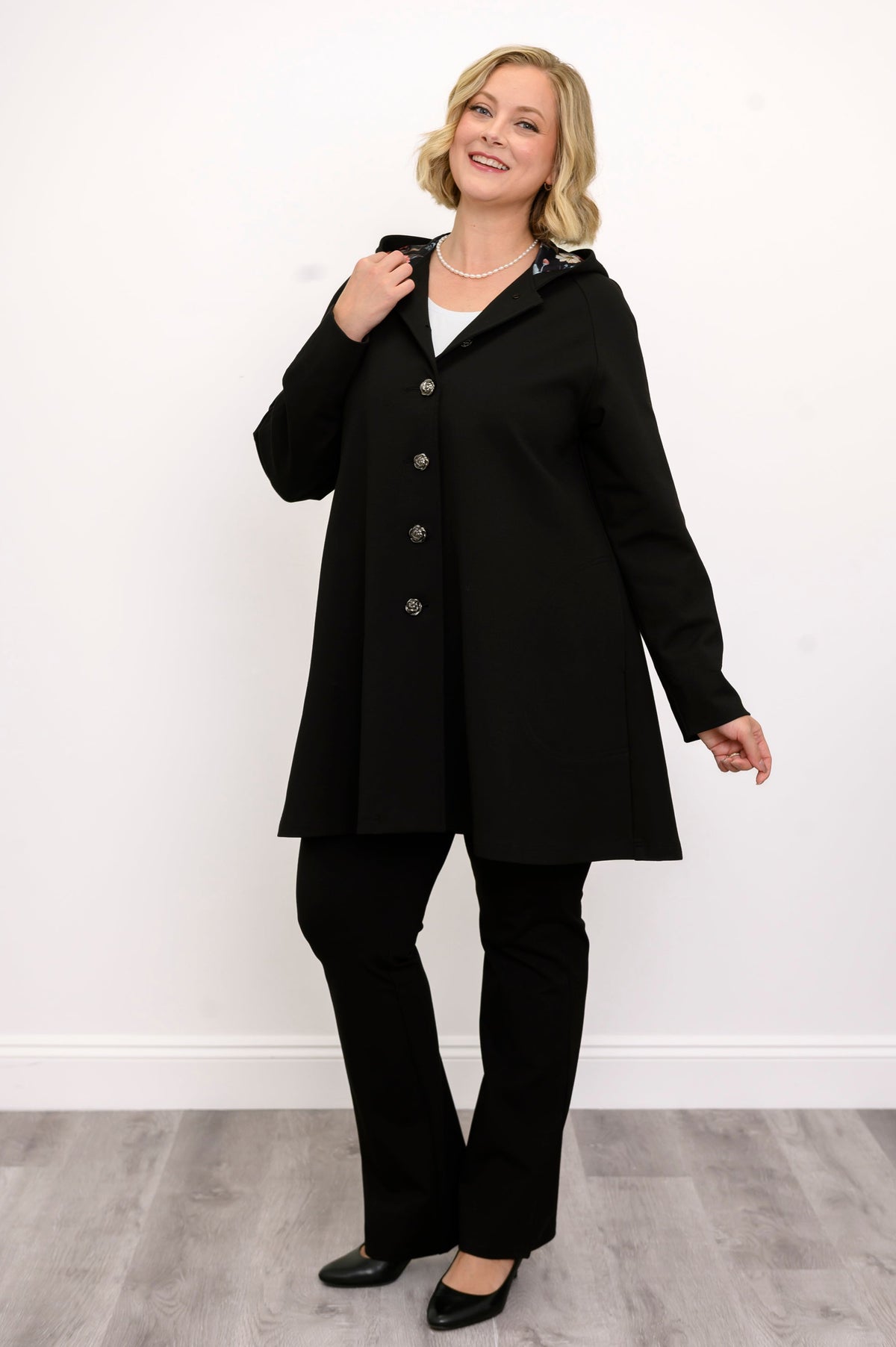 Front view of woman wearing black coat with buttons and black pants against a white background