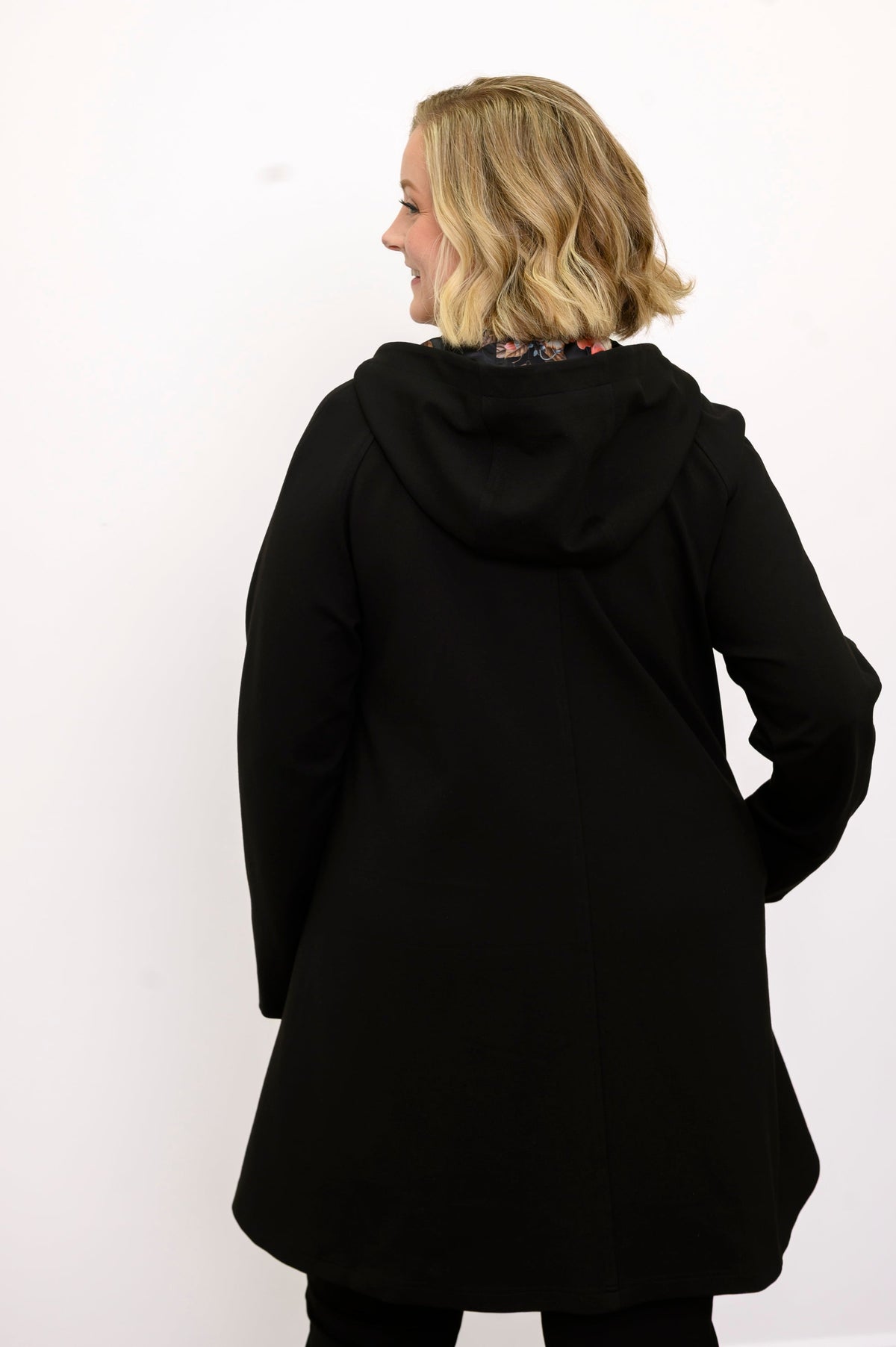 Back view of woman wearing black coat with hood, floral lining and black pants against a white background