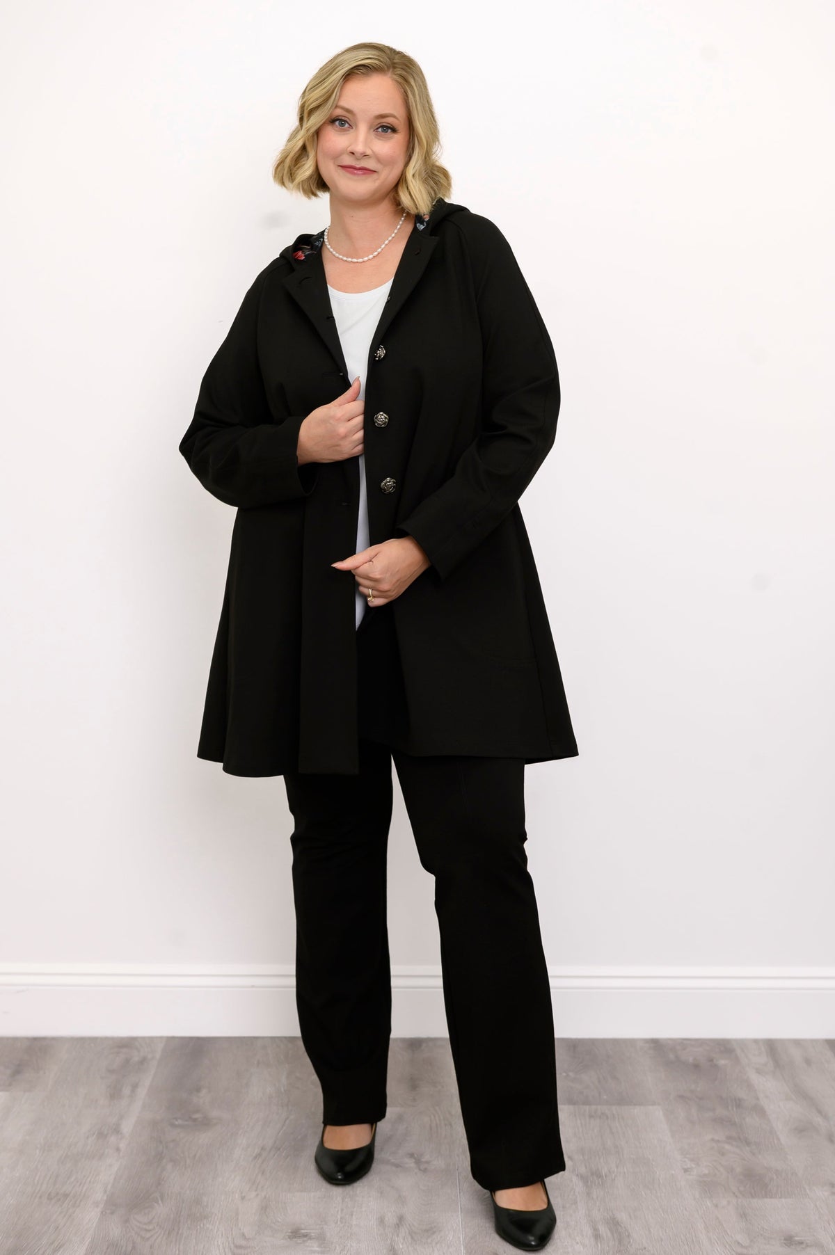 Front view of woman wearing black coat with buttons and black pants against a white background