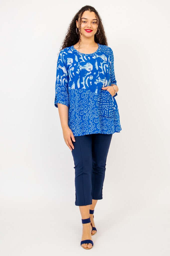 Woman standing facing forward in a blue batik Palace Blouse, hands in pocket, navy pants and sandals