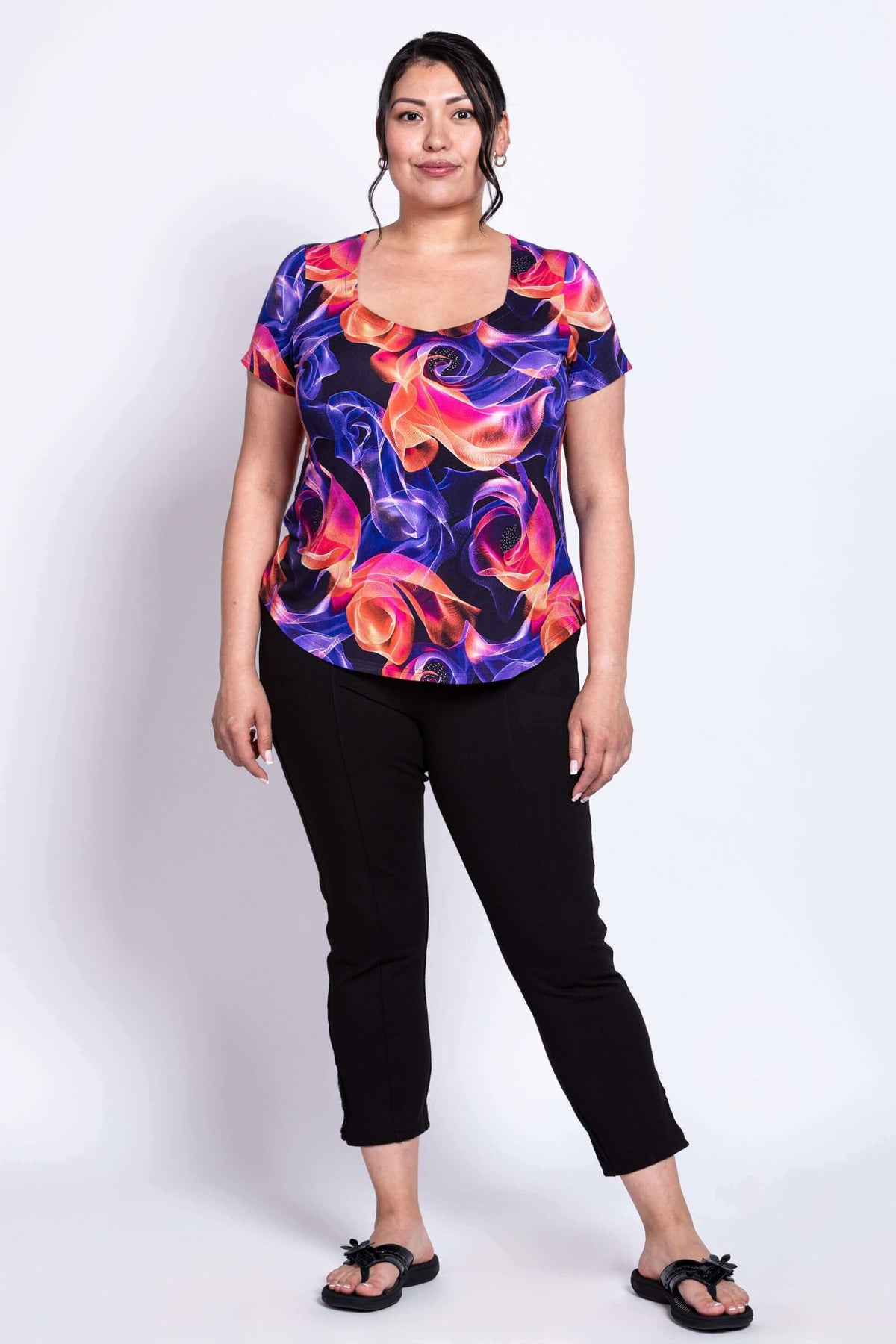 Full body front view of woman smiling and posing with her hands at her side wearing a swirl print top and black pants