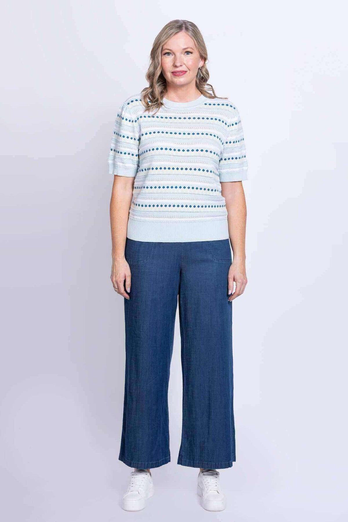 Woman in light blue eyelet stripe short sleeve sweater with wide-leg blue pants and white sneakers, front view