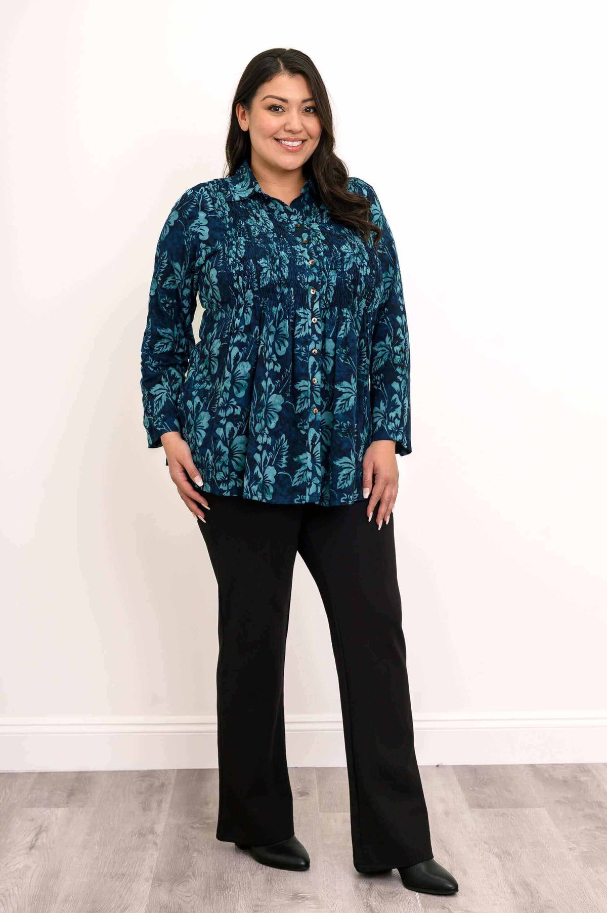 Woman in a blue floral blouse with hand in pocket and black pants, standing on wood floor