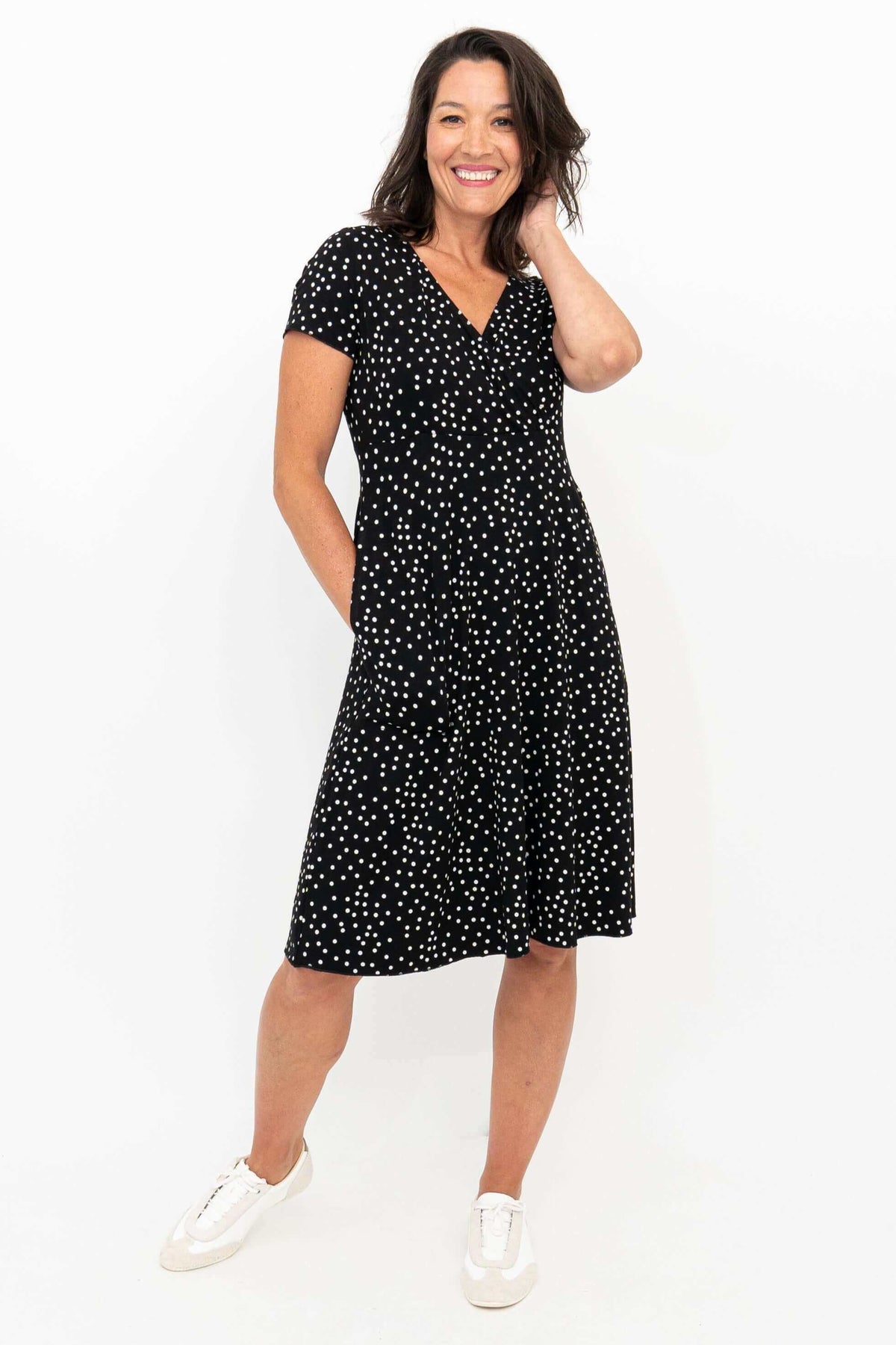 Front angled view of woman in a black and white polka dot dress with hand touching hair, smiling