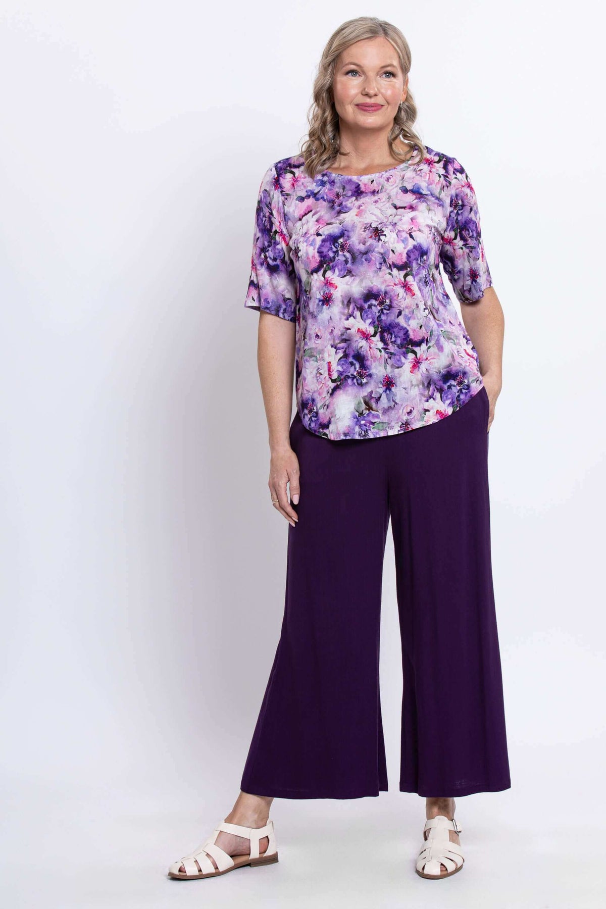 Woman standing in a purple floral blouse and pants with hands in pockets, front view