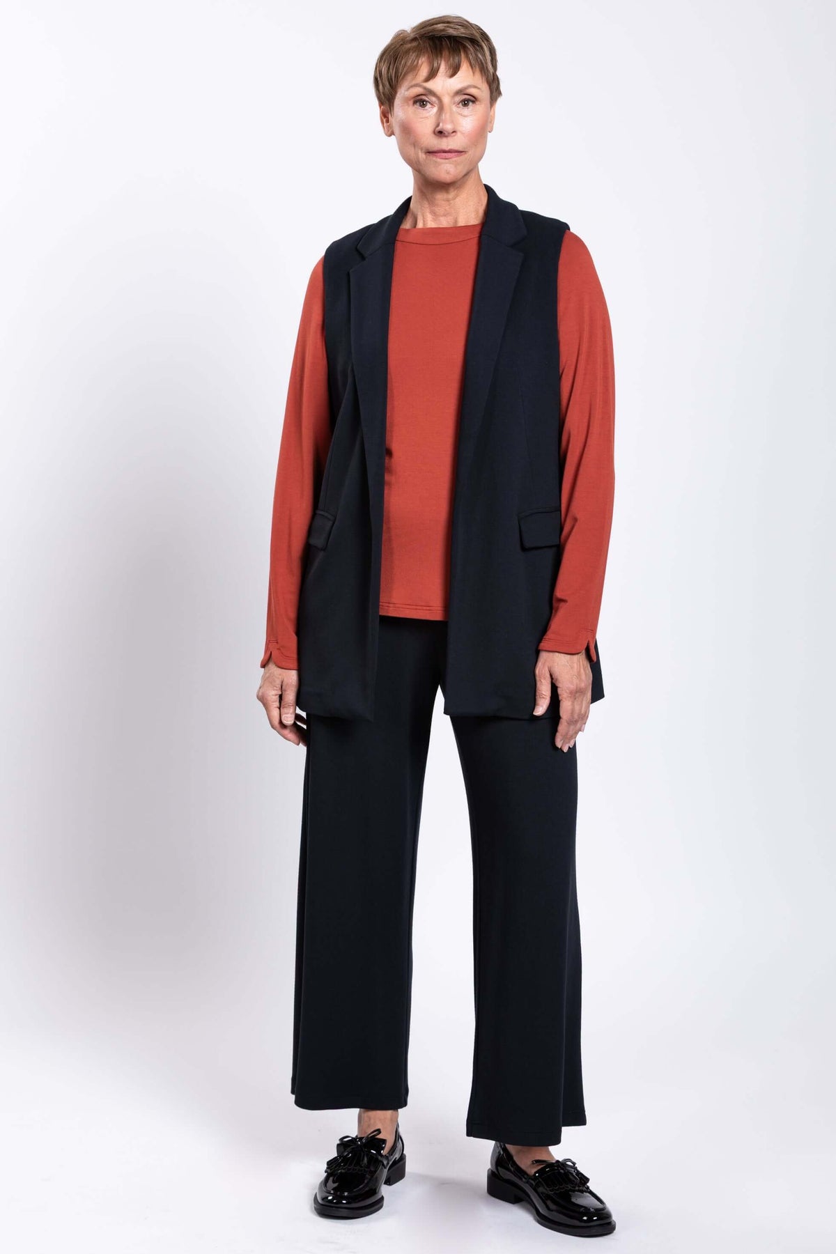 Front view of model in black open front vest, matching pants, and rust long sleeve top