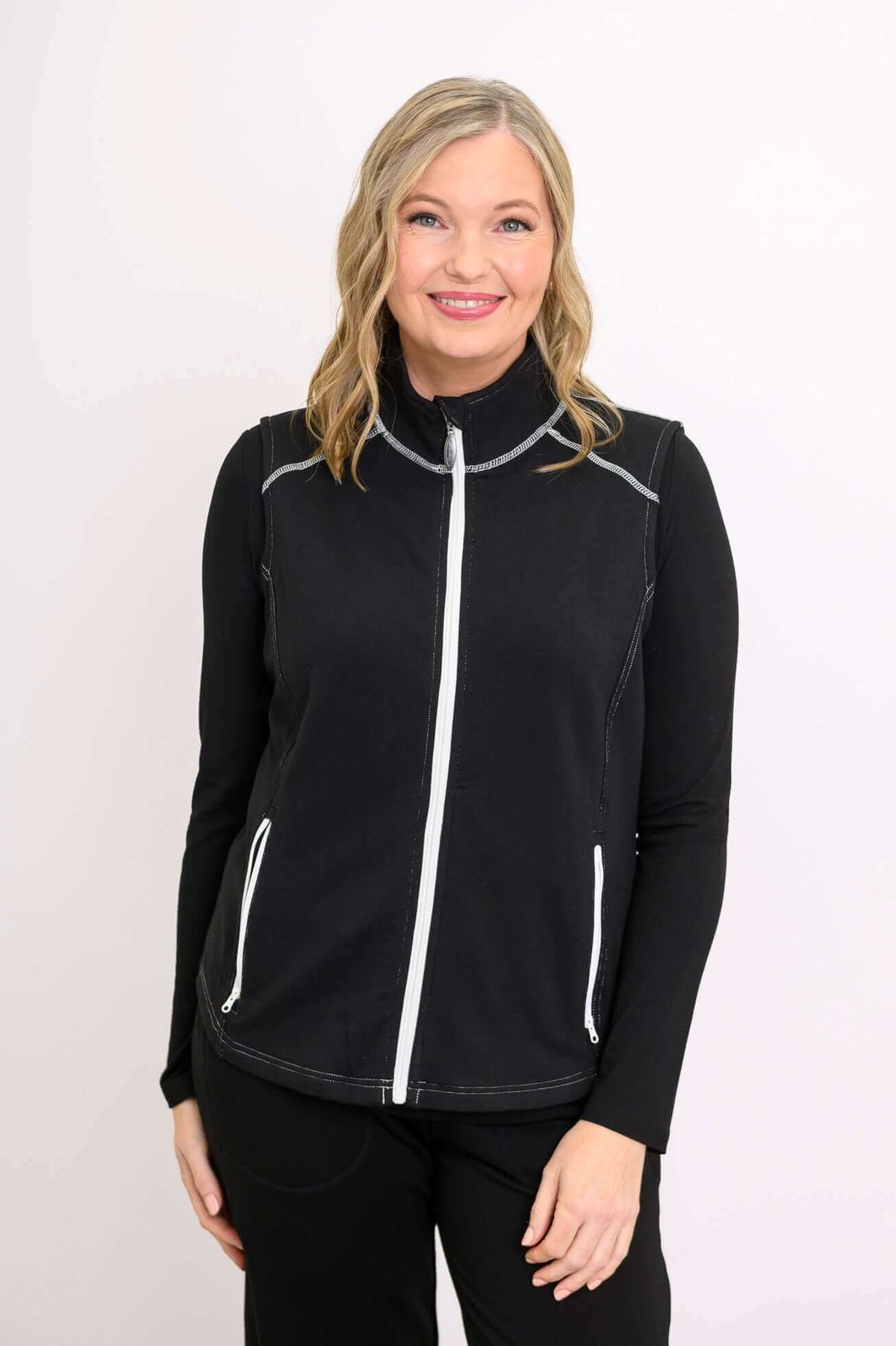 Front view of woman wearing black Hartford Utility Vest with white zipper over black long sleeve top