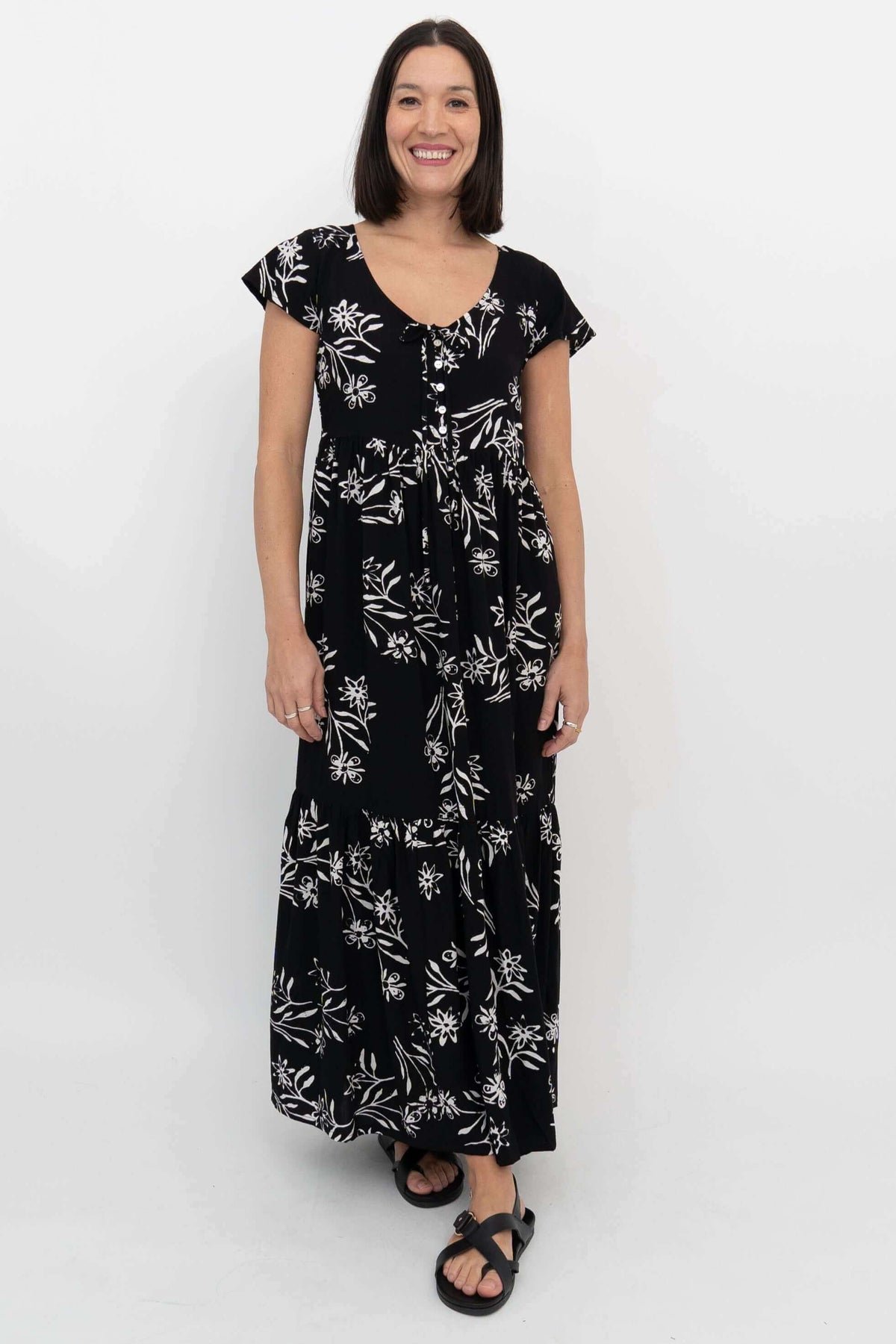 Front view of a woman wearing a black maxi dress with white floral print, flutter sleeves, and front buttons