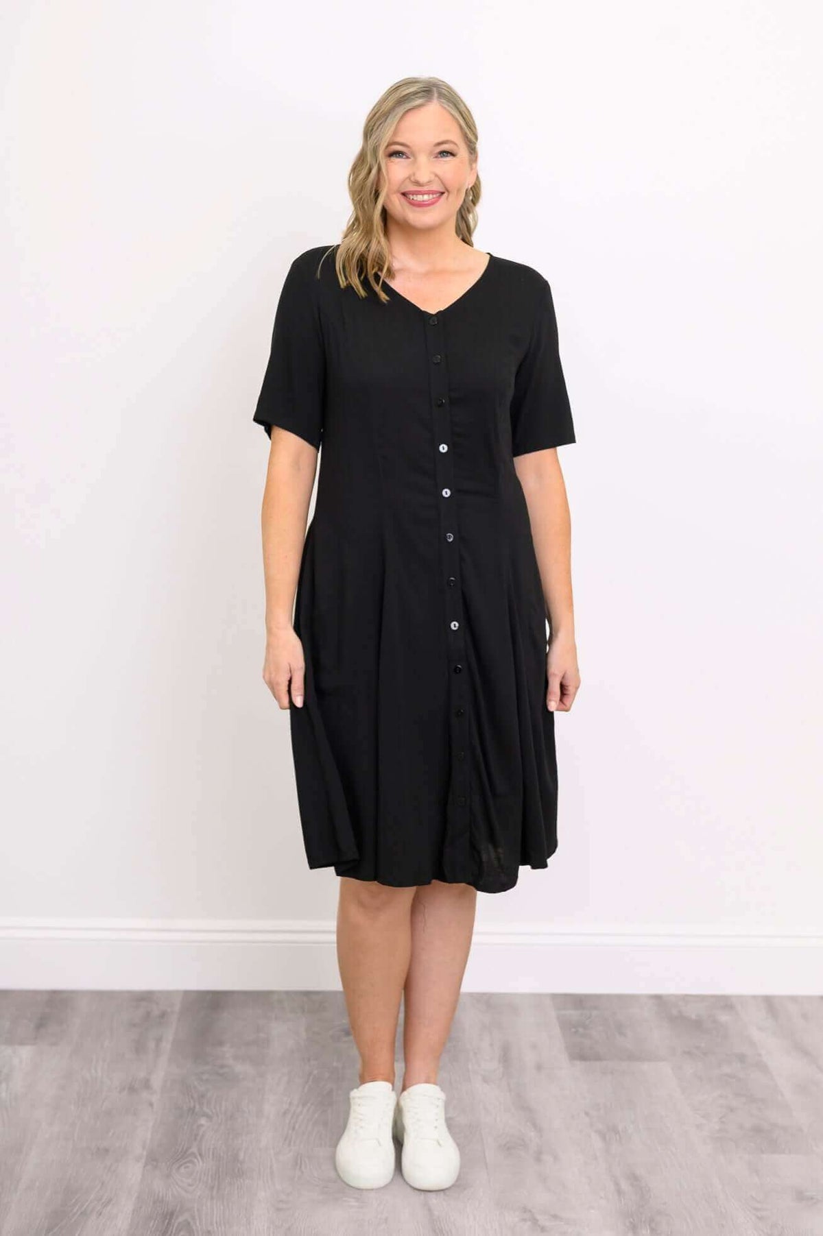 Woman smiling in a black button-up dress with white sneakers, front view