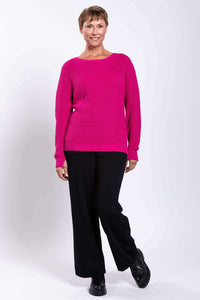 Front view of a smiling woman in a fuchsia sweater, one leg bent, with black pants