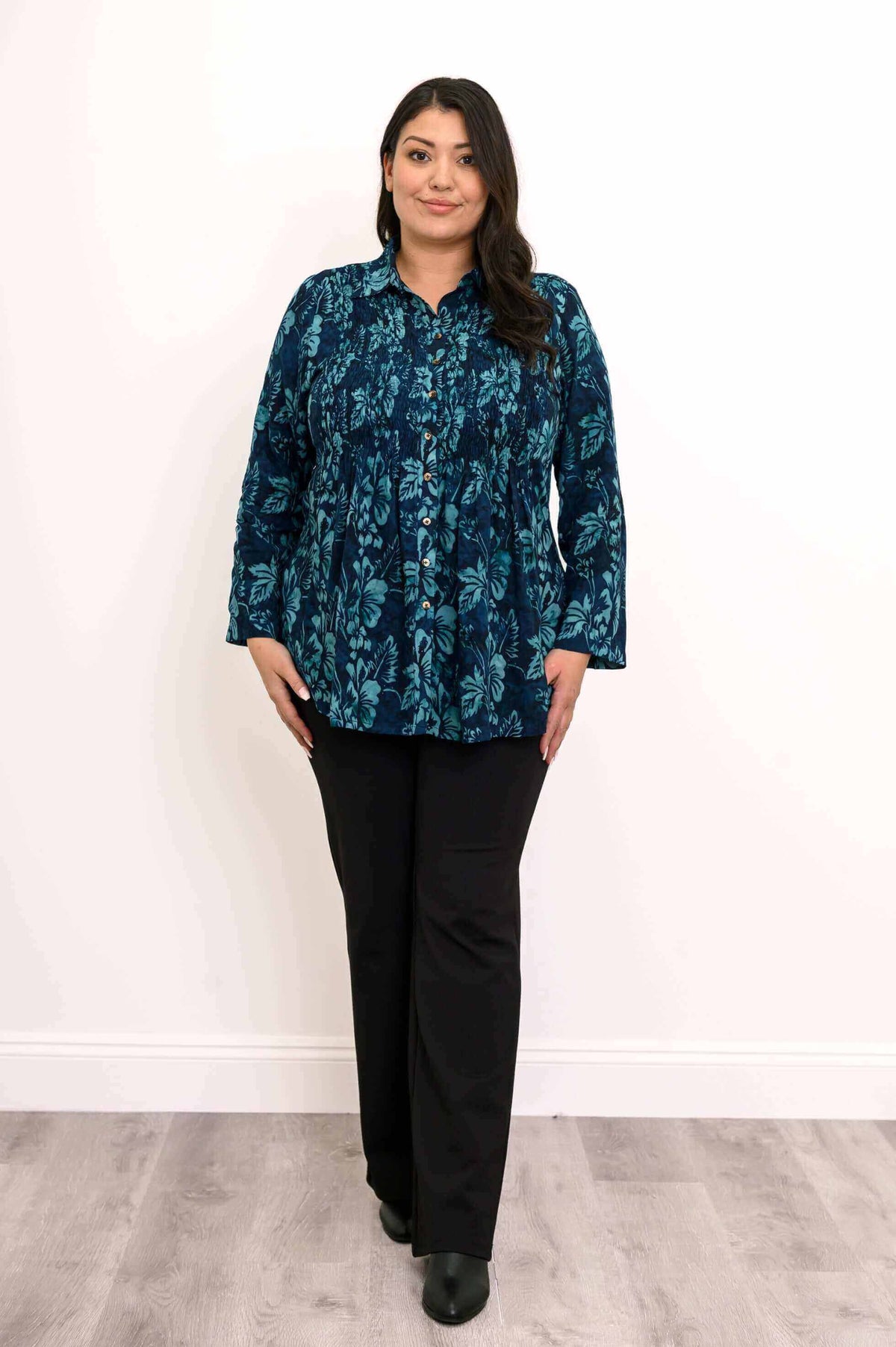 Frontal view of woman in blue floral blouse and black pants, standing on wood flooring