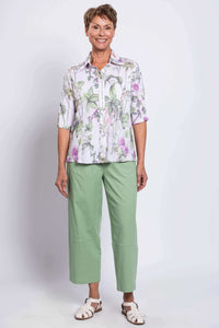 Full-body front view of woman smiling in button-up white blouse with green and pink floral pattern and green pants